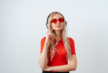 Lovely pensive blonde girl listens to music in wireless headphones, standing on a white background. The girl with headphones in red glasses and a red T-shirt thought about it.