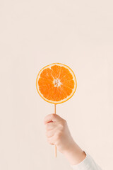 Child's hand holds a stick with an orange fruit slice on bright beige  background.  Minimal natural...