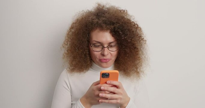 Happy cheerful woman has curly hair types text messages on smartphone looks joyfully at screen of device being addicted to modern technologies scrolls newsfeed wears turtleneck isolated on white