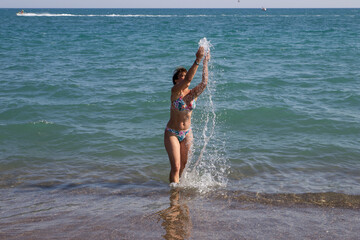 Schlanke Frau mit kurzen, dunklen Haaren und mit dem Bikini, spritzt mit dem Wasser im Meer. Sie lacht und hat Spaß im Wasser. Die Hände sind oben. Man sieht den ganzen Körper. Türkei, Fame Residence