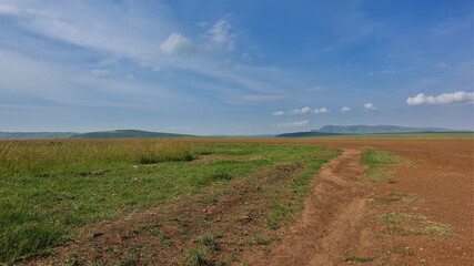 Landscape of the African savannah. The dirt road runs along the red earth. Lush green grass grows on the roadside. Silhouettes of mountains against the blue sky. Kenya. Masai Mara