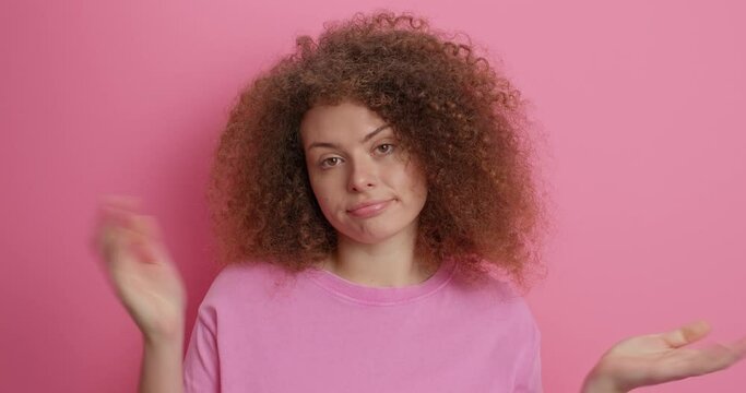 Let me think. Clueless European woman has culry hair looks away hesitant shrugs shoulders feels indecisive dressed casually raises hands up confused isolated over pink background has no idea