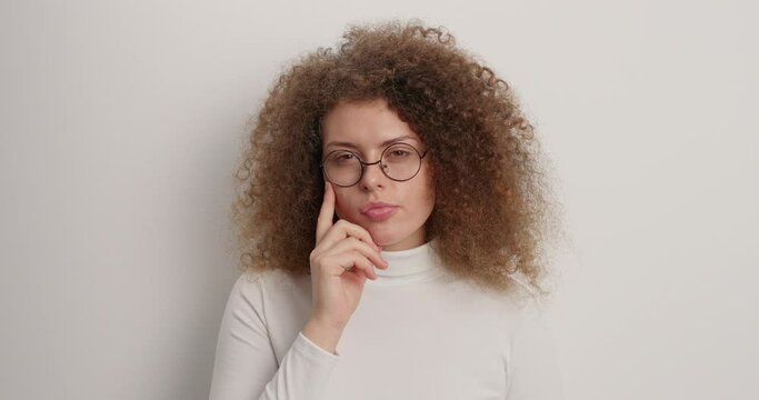 Pensive curly haired woman ponders over something with thoughtful expresion makes decision or assumptions wears casual turtleneck and round glasses isolated over white background searches solution