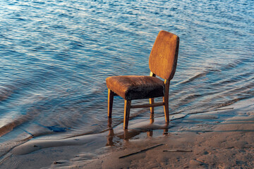 An empty soft old chair stands in the water next to the sandy bank of the river, lake. Concept.