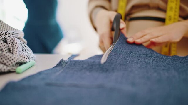 Skilled unrecognizable female tailor standing at desk, cutting denim fabric, preparing pattern for sewing, close up shot