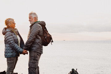 Naklejka premium Senior couple in outdoor excursion stands by the sea at sunset light. Hands in the hands, horizon over water