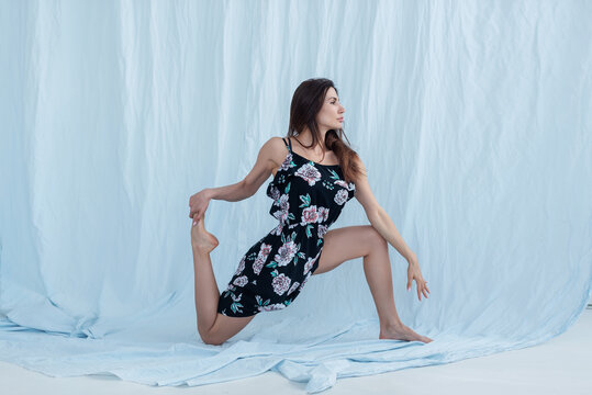 A girl with a slim figure and in a dress shows yoga asanas on a light background