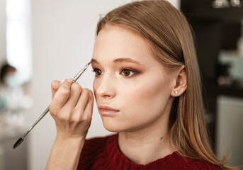 makeup artist applies eyebrow shadow on a beautiful young woman blonde model, face make up concept