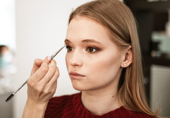 makeup artist applies eyebrow shadow on a beautiful young woman blonde model, face make up concept