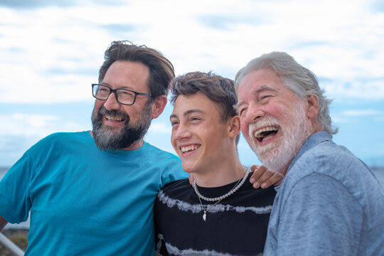 Multigenerational family group sitting outdoors by the sea, hugging each other and laughing. Grandfather, son and grandson. Horizon over the water and cloudy sky