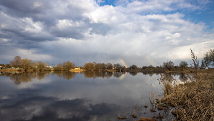 On the horizon is a village by the river. The clouds are reflected in the water. Early spring by the pond. Rural landscape with a church.