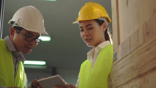 4K Young Asian woman worker use tablet and man engineer supervisor stand to talking check product box shelf in big warehouse factory store which smile and felling happy, logistic and transportation