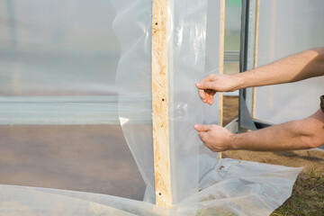 Young adult man hands stretching new polythene film on greenhouse metal profile carcass. Closeup. Side view. Preparation for garden season in spring.