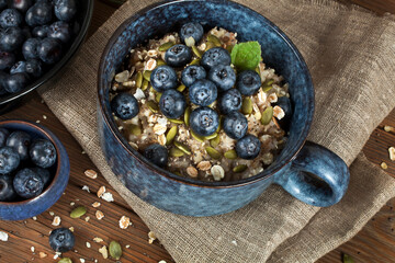 Oatmeal porridge with blueberries and seeds in a blue ceramic bowl and a glass jar of oat flakes on a wooden background. Healthy eating concept.