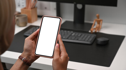Close up view young woman holding mock up mobile phone with white screen while sitting at her workplace.