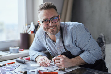 Male artist designer in glasses, shirt and black apron sitting on a studio background. Portrait of...