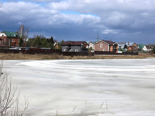 country pond, covered with a crust of spring ice with cozy houses in the distance