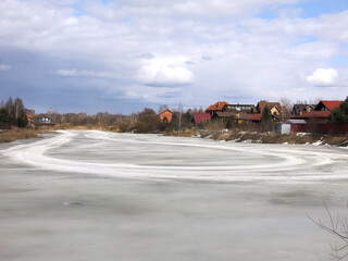 Obraz premium country pond, covered with a crust of spring ice with cozy houses in the distance