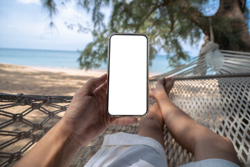 Hand holding mock up mobile with white sceen while laying on hammock swing between trees on the beach.