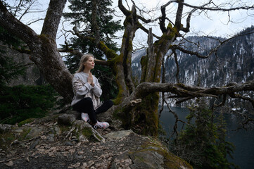 Fototapeta na wymiar Girl sits on a tree and meditates on blue lake background. Lake Ritsa in Abkhazia