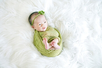 opened eyes Newborn wrapped in a green cloth with a green flower headband showing little feet lying on a soft white flokati - Powered by Adobe