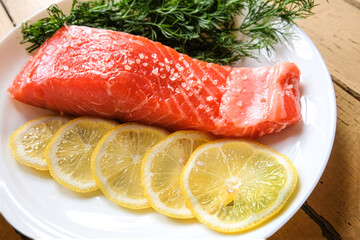 A piece of fresh trout fillet with lemon and dill greens on a white plate on a wooden table waiting for cooking. Restaurant menu. Dietary seafood. Keto and paleo diets. The concept of healthy eating.