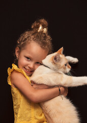 Obraz premium A little happy girl holds her cat in her arms. Portrait on a black background