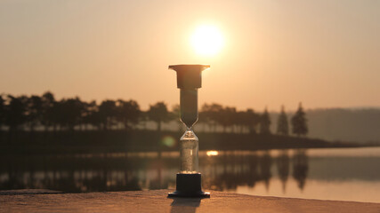 Obraz na płótnie Canvas Hourglass outdoors during dawn. Start of a new cycle of life, new day. Value of time in life, an eternity. Hourglass on the background of the lake and the rising sun.