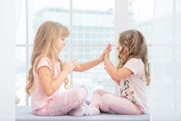Cute little sisters sitting on windowsill at home playing with each other