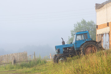 Old blue agricultural tractor near a brick building. Morning fog. Summer rural landscape. Agricultural machinery on the outskirts of the village.