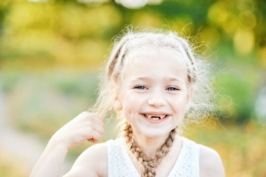 Funny girl with hair plaits. Kid expressing emotions. Close up portrait of beautiful joyful blonde Caucasian girl smiling demonstrating white teeth without two front teeth. Cheerful child.