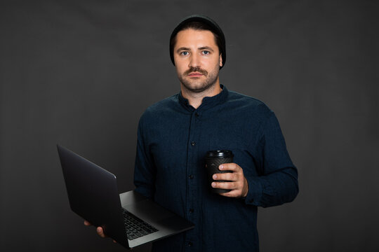 Portrait of young hipster moustached man wearing hat posing on isotaled grey background holding laptop and coffee cup. Concept of freelance work, IT professions, remote study, online education courses