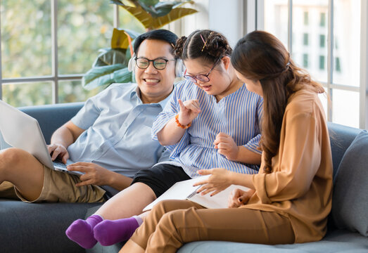 Happy Asian Family Father Mother And Handicapped Down Syndrome Child Sitting On A Couch Relaxedly In A Living Room At Home. The Daughter Learning How To Count A Number