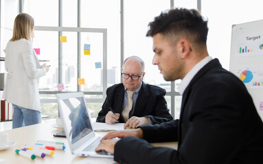 Focus on Business manager wearing eyeglass writing on paper and staff sitting typing and seeing on laptop near colour pens. Woman staff holding pen and seeing sticky paper snap on mirror at office