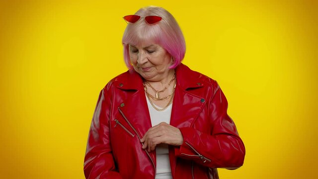 Cheerful lovely senior old woman rocker with pink hair in red leather jacket posing isolated on yellow studio wall background. People lifestyle. Elderly stylish smiling grandmother looking at camera