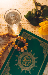 The Al Quran with written arabic calligraphy meaning of "Holy Quran", rosary (beards), glass of water and a rose on the table during suhoor or iftar in the month of Ramadan. Ramadan concept.