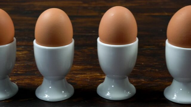 Panning shot along a row of boiled eggs in white egg cups to one egg smiling at a broken egg