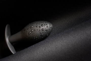 A wet black butt plug lies on a dark surface. Moisture or lubricant drops. Sex toy and pleasure. Diagonal. Close-up. - obrazy, fototapety, plakaty