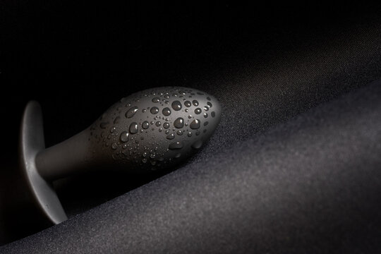 A wet black butt plug lies on a dark surface. Moisture or lubricant drops. Sex toy and pleasure. Diagonal. Close-up.