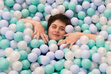 Fototapeta na wymiar Portrait of a young brunette woman immersed in a pool with plastic balls. Mom in the children's entertainment center has fun while the child plays. 