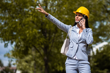 Gorgeous caucasian female architect in gray suit and with protective helmet on, standing at construction site and using smart phone.