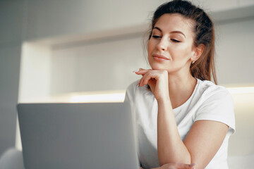 A young woman is working on a laptop in an apartment. Online video communication with relatives. A brunette with a ponytail makes an order in an online store with home delivery.