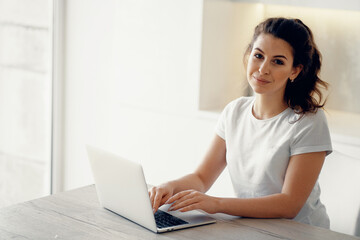 Fototapeta premium A brunette of European appearance. Does work on a new project remotely. Orders online purchases in the apartment. Portrait of a woman sitting at home and typing on a computer text.
