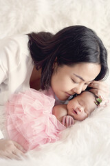 Obraz na płótnie Canvas Asian mom kisses her baby with love. The child smile with happiness. Sleeping newborn is wearing a pink tutu showing small hands with white blur background