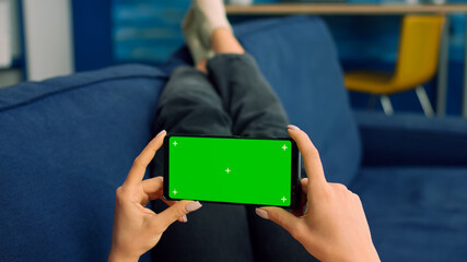 Business woman lying on sofa holding isolated phone with mock up green screen chroma key during social media media communication. Caucasian female browsing on internet watching online videos
