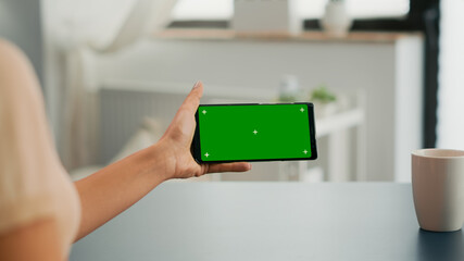 Freelancer holding smartphone with mock up green screen chroma key in horizontal position. Business woman searching online information using isolated device sitting on office desk