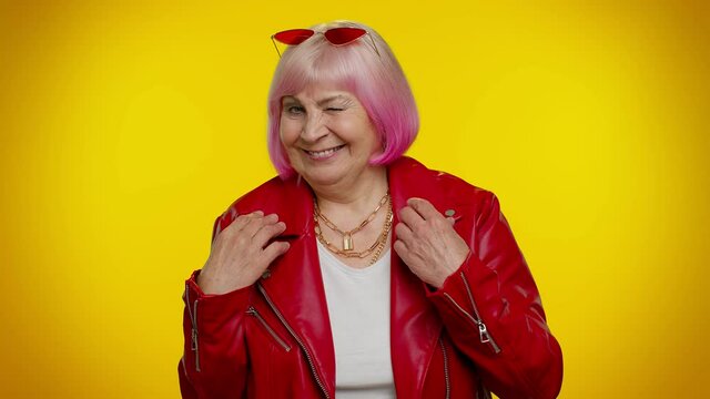 Playful happy elderly rocker granny woman in jacket blinking eye, looking at camera with toothy smile, winking and flirting, expressing optimism. Senior mature old emo grandmother on yellow background