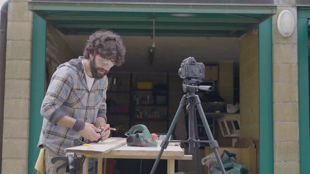 Young male craftsman showing how to work, take measurement on wood furnitures self-recording video on reflex camera, outside garage shop. online course learning, distant teacher icons. Slow-motion