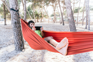 Hispanic man lying in a hammock and playing video games at Palmanova beach. Palma de Mallorca, Spain (Perfect for Copyspace)	