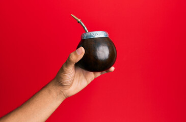Hand of hispanic man holding cup of mate infusion beverage over isolated red background.